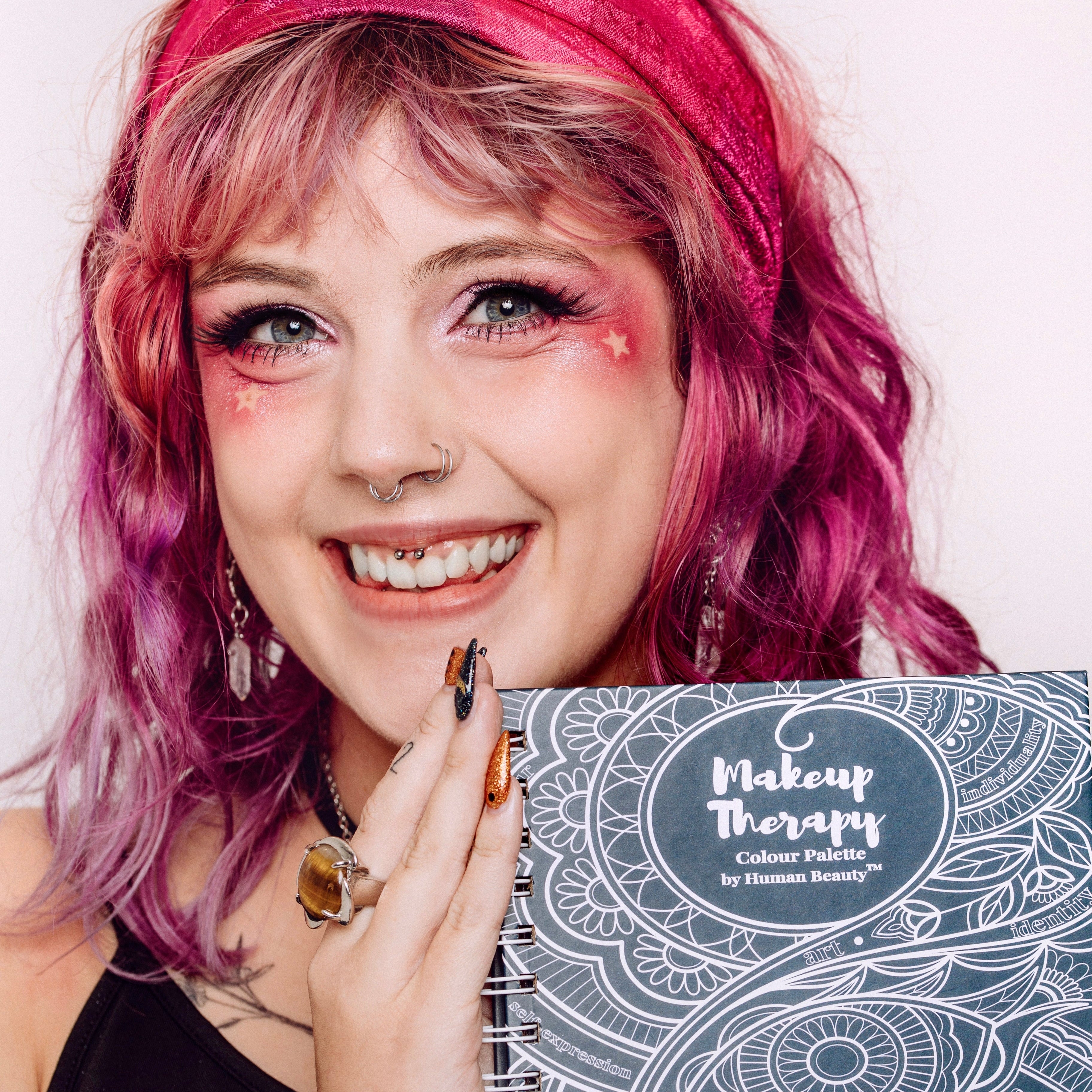 Woman with pink hair and scarf holds Makeup Therapy Palette book, highlighting the palette&