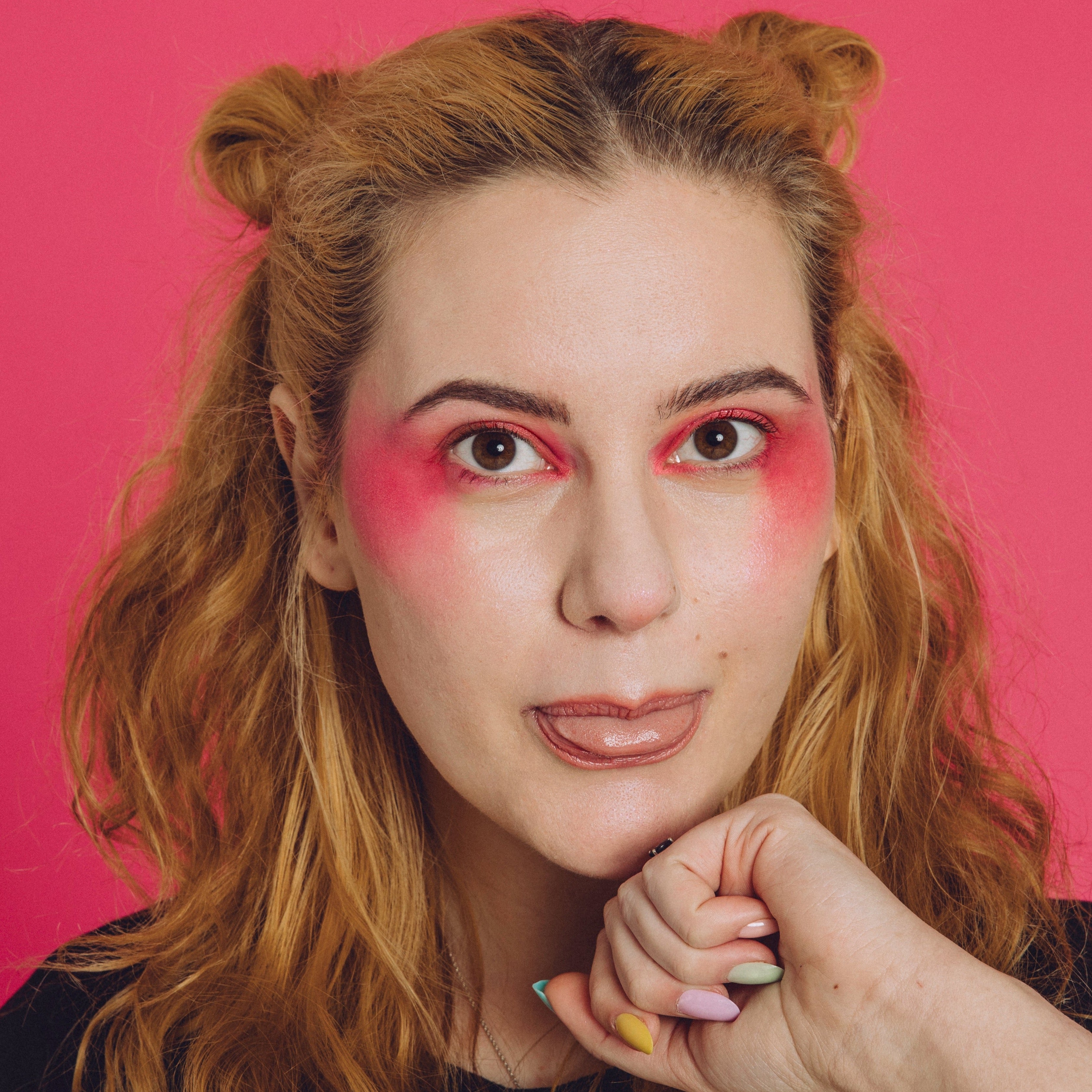 White woman with blonde woman wears a bright pink eyeshadow look across her cheeks and eyelids. She sticks her tongue out.