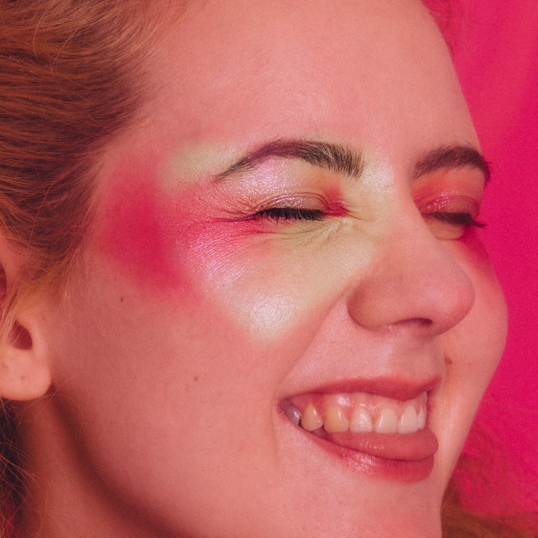 White woman smiles with her tongue out. She is wearing a pink shimmer eyemakeup look which extends to her cheeks.