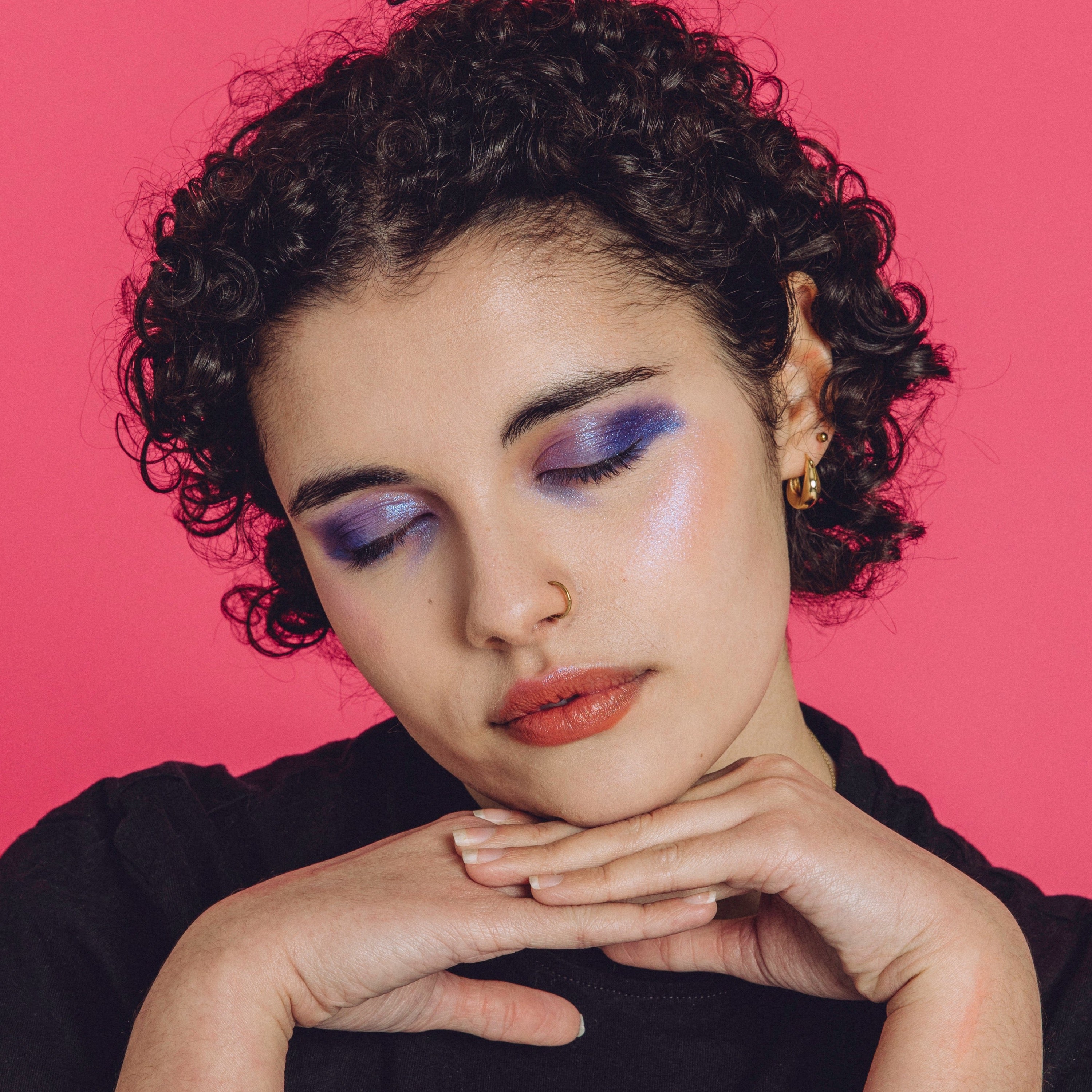 Woman with black curly hair wearing a shimmery purple and blue eyeshadow look and closing her eyes.