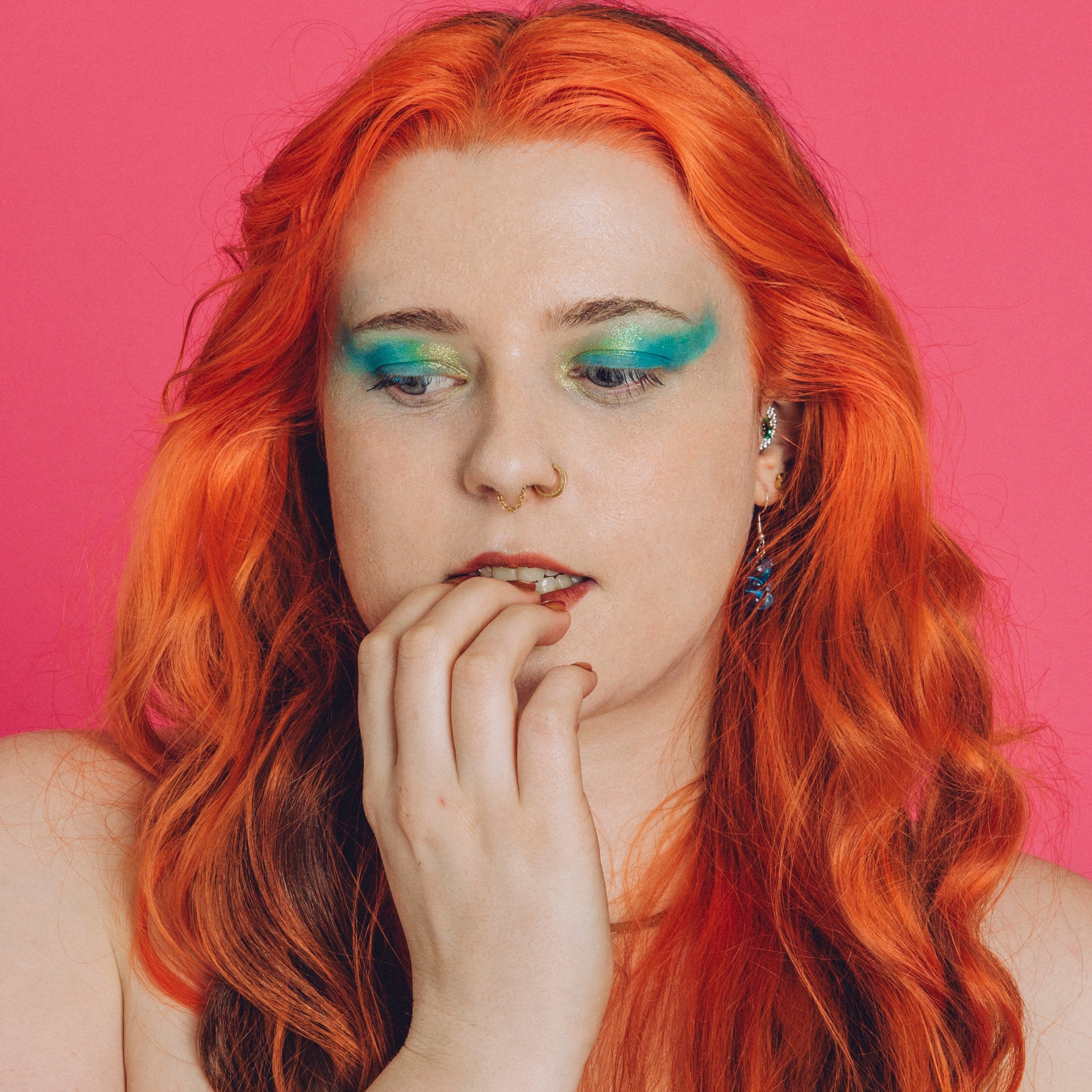 White woman with orange hair wears a shimmery gold and green eyeshadow look and bites her nails, looking anxious.