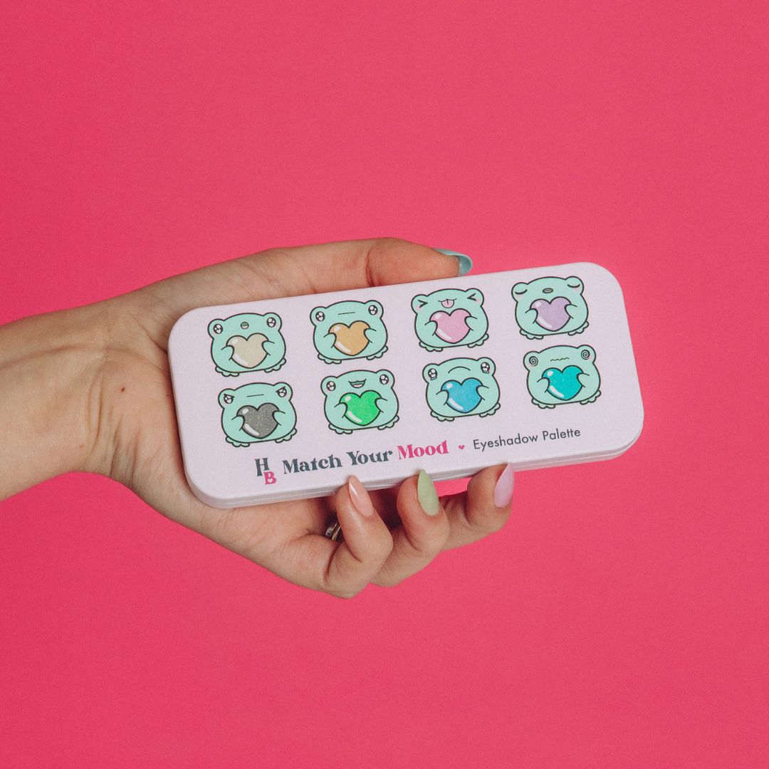 the match your mood palette held closed by a hand with fair skin on a pink background. it is light purple coloured with the Human Beauty logo and the name: Match Your Mood eyeshadow palette, as well as 8 frogs designed by Fuzziesartdesign embodying different moods: surprised, hungry, silly, tired, angry, happy, sad and anxious