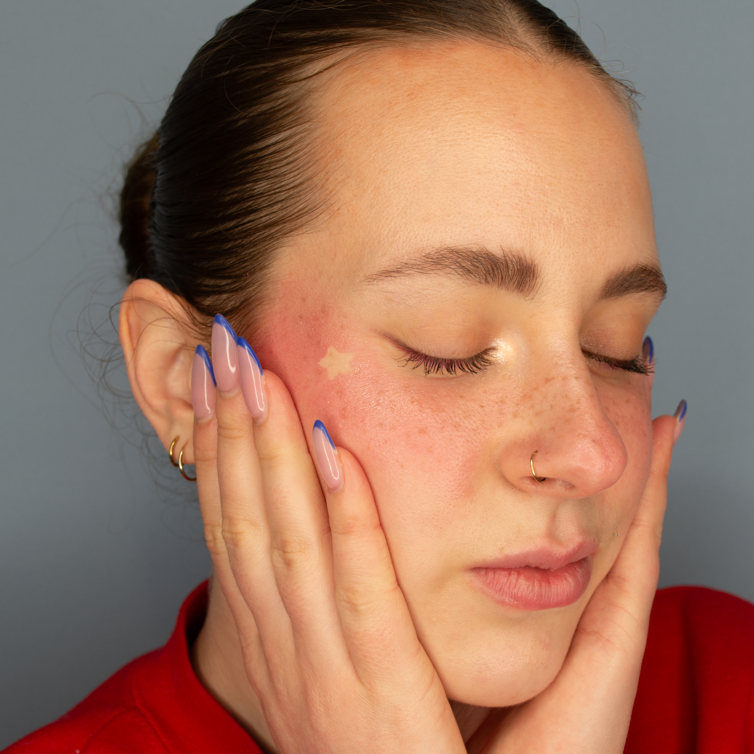 Close up picture of a model wearing the Dare To Blush heart-shaped blush stick in shade Courageous Crimson on her cheeks. She has fair skin and her hair is in a sleek bun. She is holding her face in her hands