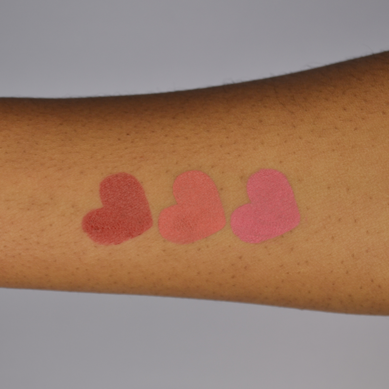 Swatches of the Dare To Blush Opulent Berry bundle: shades Courageous Crimson, Resilient Rosé and Bold Berry in heart shapes on dark skin