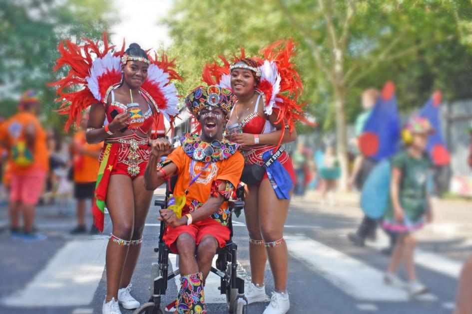 Three Harrow Mencap ambassadors at Notting Hill Carnival. Featuring a person in their wheelchair, with a big smile and wearing a traditional carnival headdress. Stood eitherside are two ambassadors wearing red and white feathered carnival outfits. 