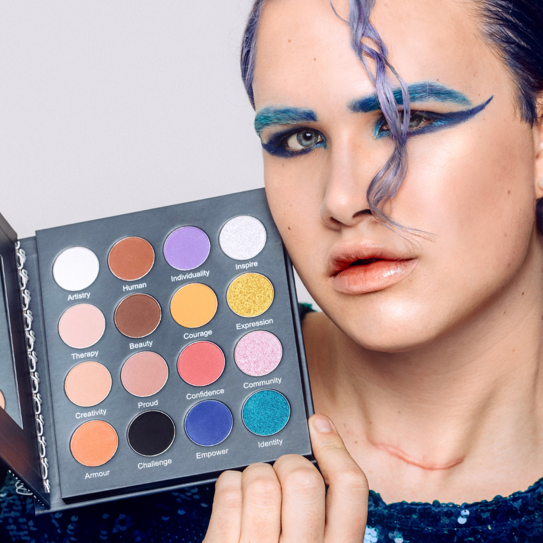 Man wearing a blue winged eye makeup look with blue eyebrows holds open the Makeup Therapy palette.