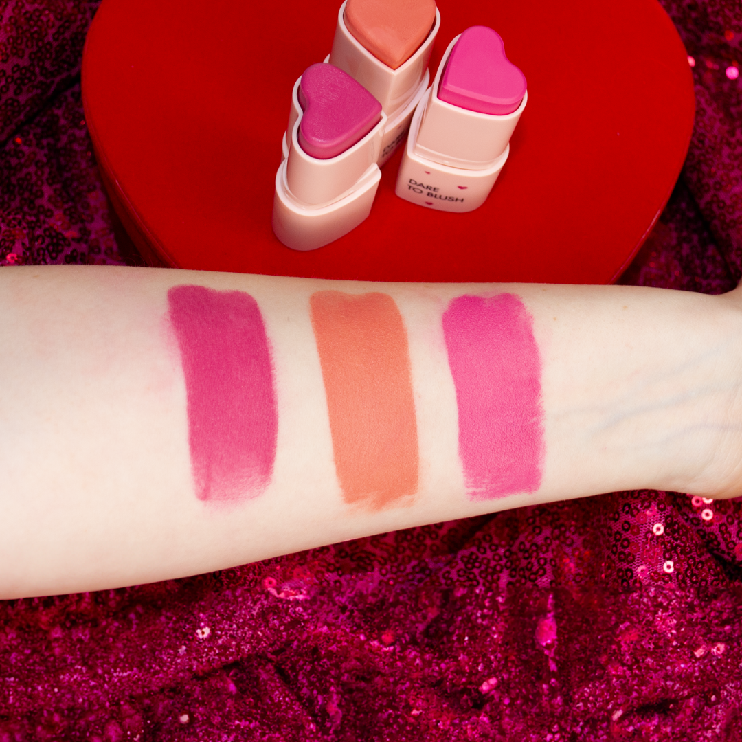 swatches for the happy hearts blush trio on fair skin: shades confident coral, bold berry and protagonist pink on a valentines day decor with sparkly hot pink fabric and a big red heart box