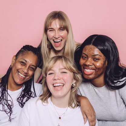 A group of women smile at the camera. Features two black women, one with braids and one with black hair, and two white women, one with longer hair.