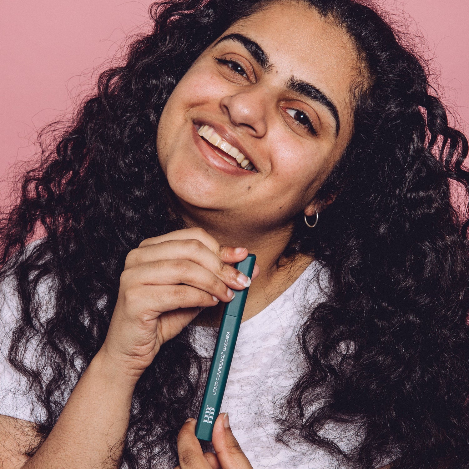 South Asian woman with long curly black hair smiles whilst holding the Liquid Confidence Mascara.