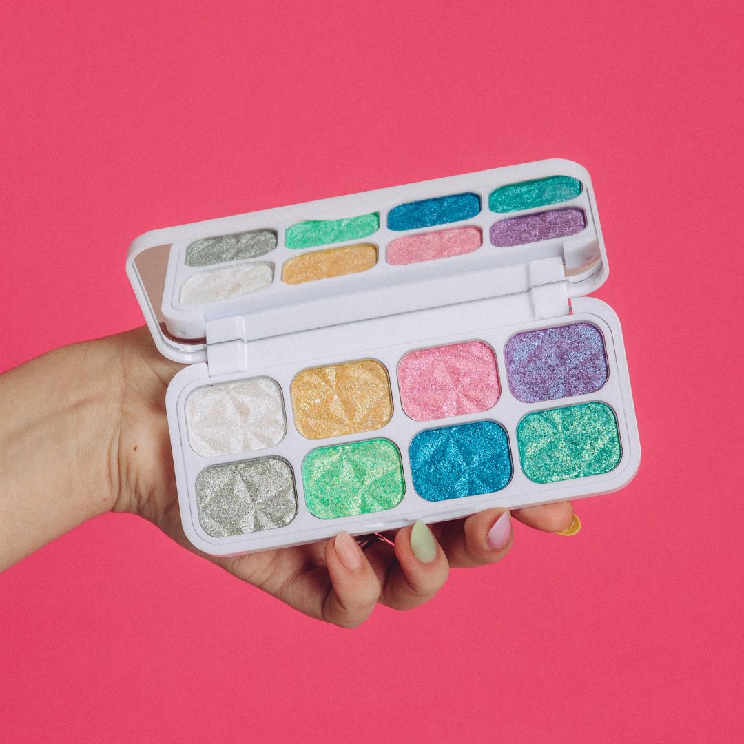 the match your mood palette held open by a hand with fair skin open on a pink background. it is light coloured with eight pans of reflective shadows in colours: white, yellow, pink, purple, grey, light green, blue and teal