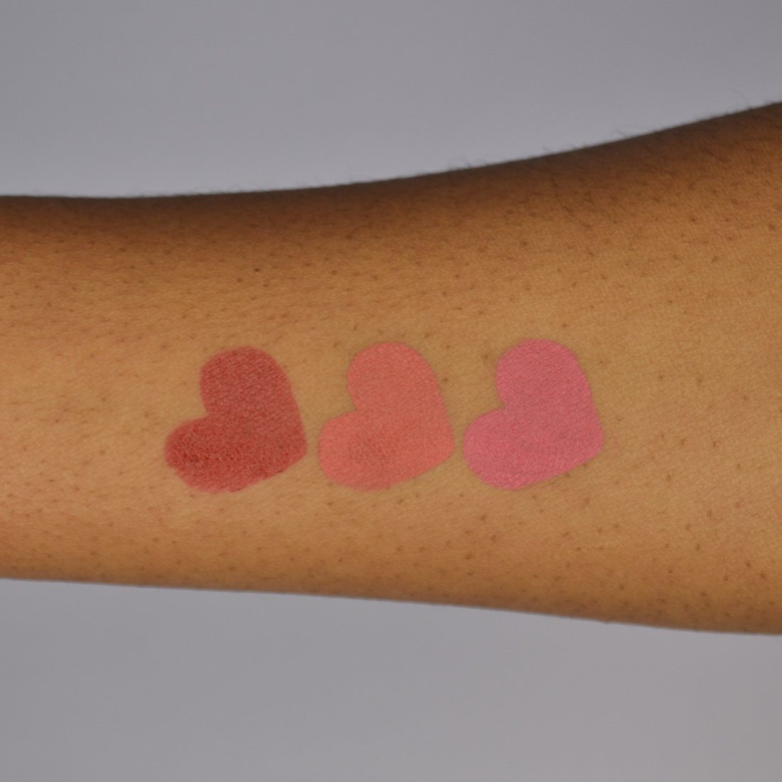 Swatches of the Dare To Blush Opulent Berry bundle: shades Courageous Crimson, Resilient Rosé and Bold Berry in heart shapes on dark skin
